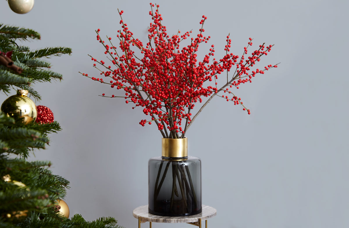 Genuine Gardenia Christmas red Ilex berries in a glass vase beside a decorated tree